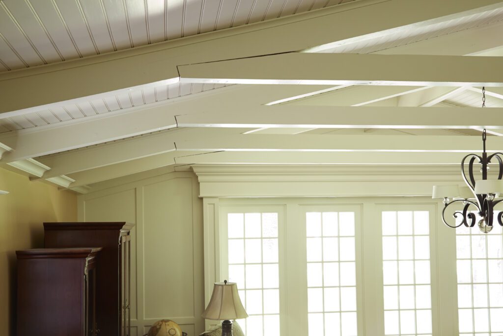 A bright room with large French doors letting in natural light. The ceiling features white exposed beams, a testament to the skill of a master remodeler. Part of a chandelier, a wooden cabinet, and a table lamp are visible in the decor. The room has a cozy, airy atmosphere.