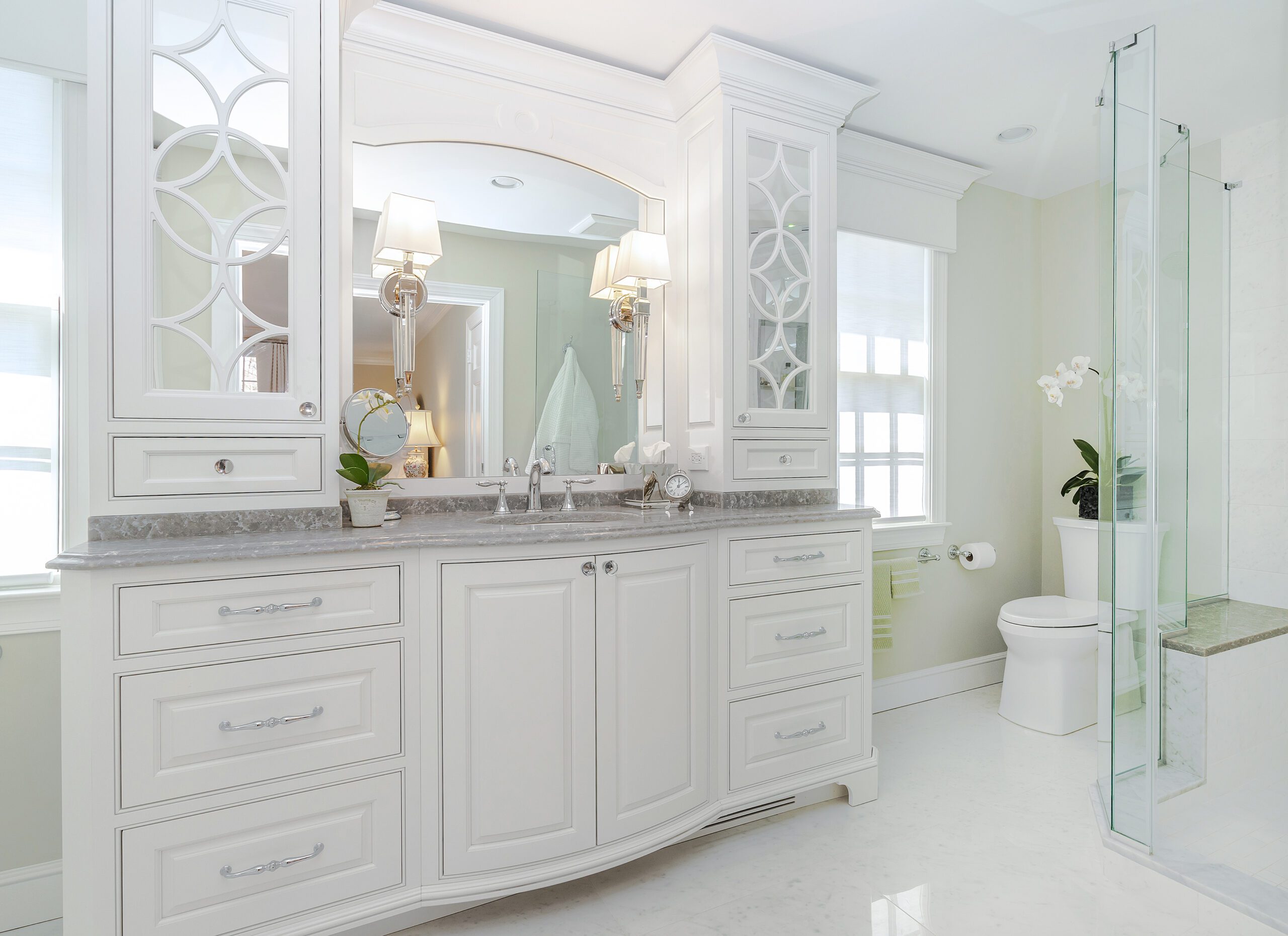 A spacious, elegant bathroom with white cabinetry and marble countertops. The vanity features double sinks, ornate mirrors, and wall sconces. A glass shower enclosure and a white toilet are on the right. The room is bright, with natural light filtering through window blinds.