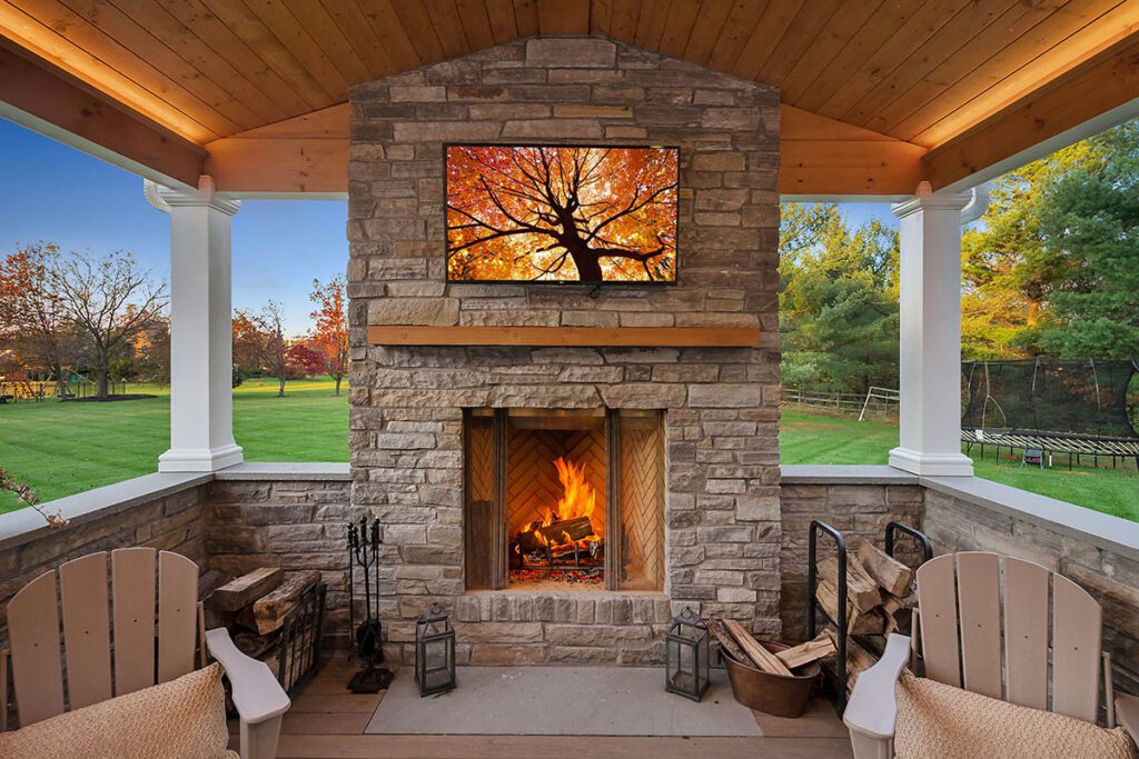 A cozy outdoor patio with a stone fireplace featuring a roaring fire. Above the mantel, there's a TV displaying an image of a tree with autumn foliage. The space is flanked by white columns, and there are two wooden chairs and stacks of firewood on either side.