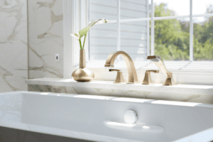 A luxurious bathroom with a white marble bathtub and gold fixtures beneath a large window. Sunlight streams in, illuminating a gold vase with a green leaf on the window ledge. The backdrop features matching marble walls—a perfect home remodeling inspiration.