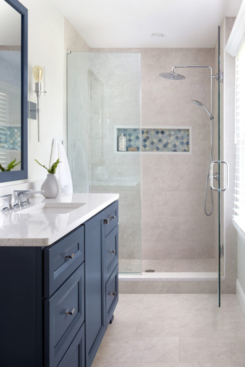 A modern bathroom, perfect for home remodeling, features a glass-enclosed shower with a rainfall showerhead and decorative tile inset. The vanity boasts a white countertop, blue cabinets, greenery in a vase, and a large mirrored cabinet. Natural light floods the well-lit space.