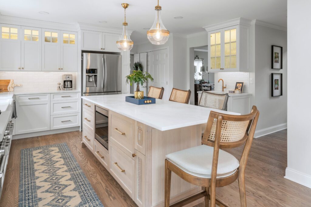 A modern kitchen featuring a large island with a white countertop and wooden base, surrounded by wooden chairs with woven backs. Above the island are two pendant lights. The background includes white cabinetry, stainless steel appliances, and a small potted plant.