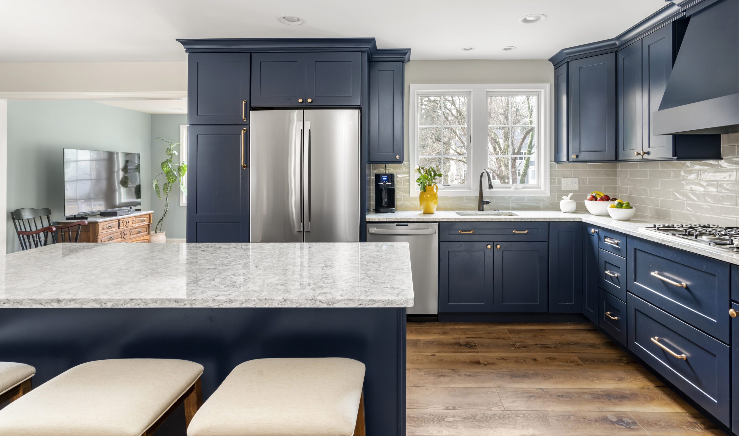 Modern kitchen with navy blue cabinets, stainless steel appliances, and a marble countertop island. Light streaming through large windows illuminates the hardwood floors and white subway tile backsplash. Three beige cushioned stools line the front of the island, showcasing impeccable design build remodeling.