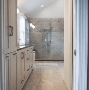 A modern bathroom in a master suite remodel, with light gray tiled floors and walls, features a glass-enclosed shower area with a large showerhead. On the left, there is a white vanity with a dark countertop and double sinks. A window with blinds allows natural light to fill the room.
