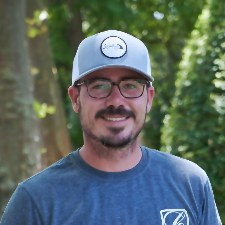A man with a beard and mustache, wearing eyeglasses, a grey baseball cap with an emblem, and a blue t-shirt, stands outdoors. Background includes green foliage and sunlight filtering through trees. He is about to smile at the camera.