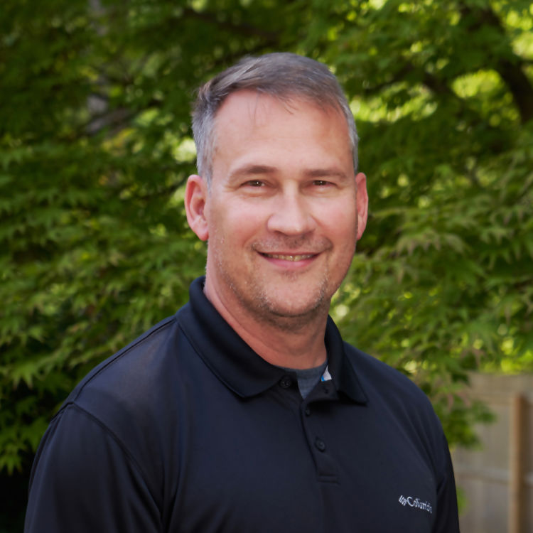 A man with short hair is smiling at the camera. He is wearing a black polo shirt with a light emblem on the chest. The background, filled with lush green foliage, suggests he is outdoors on a sunny day, perhaps enjoying everything about nature.