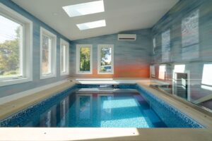 An indoor swimming pool with blue mosaic tiles, surrounded by beige deck with floor skylight windows. The room has large windows on the left wall letting in natural light and colorful, gradient sunset wall mural. An air conditioning unit is mounted on back wall.