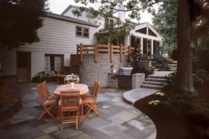 A spacious outdoor patio with a wooden dining set for six, situated on a stone-tiled surface. A modern grill is set against a raised stone wall with wooden railing. Nearby, stone steps lead up to a higher terrace, and the area is surrounded by lush greenery.