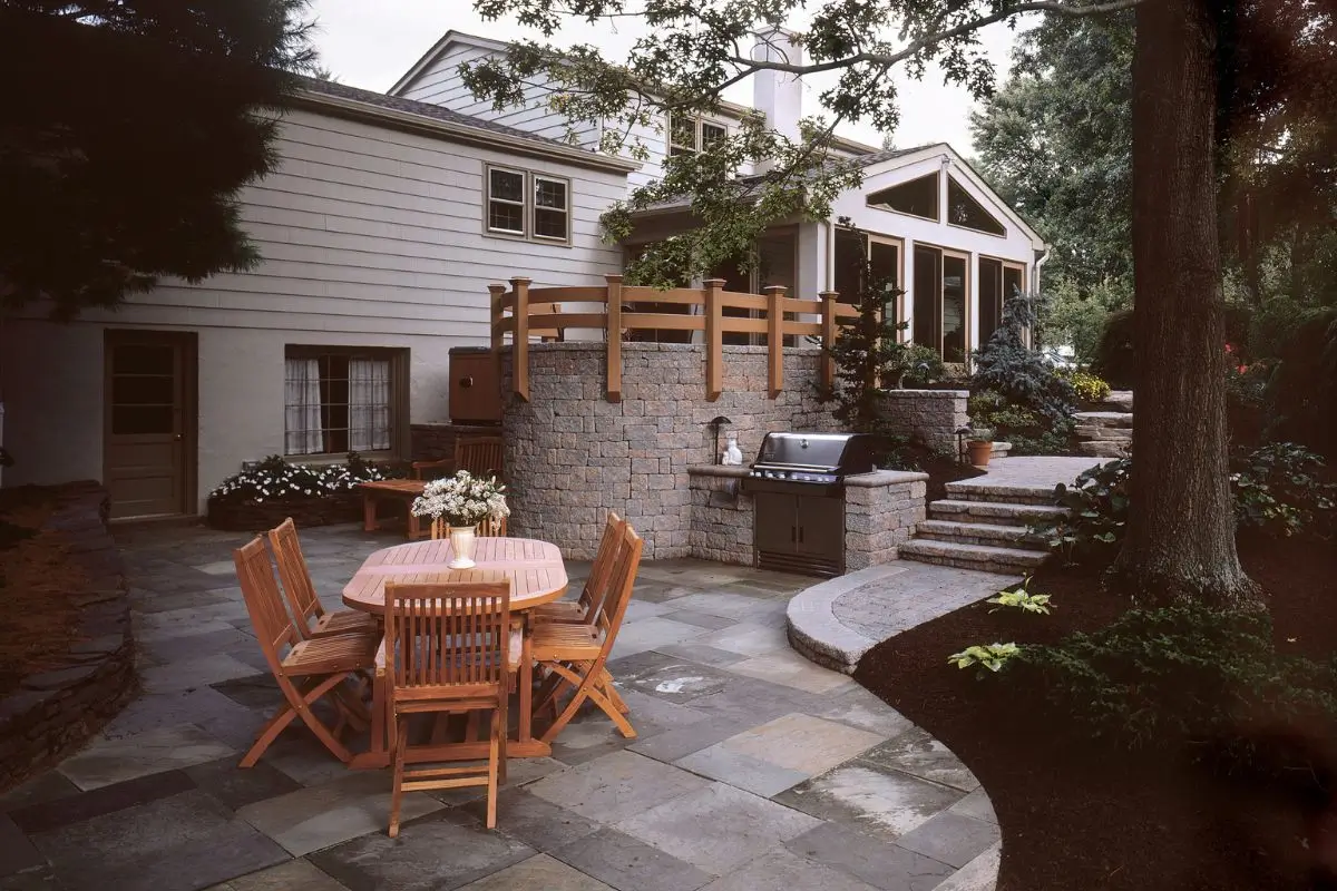 A spacious outdoor patio with a wooden dining set for six, situated on a stone-tiled surface. A modern grill is set against a raised stone wall with wooden railing. Nearby, stone steps lead up to a higher terrace, and the area is surrounded by lush greenery.