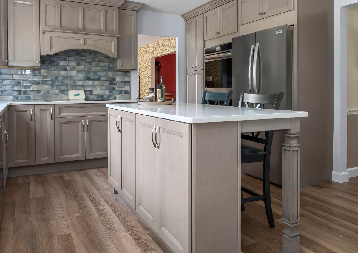 A modern kitchen renovation with taupe cabinetry, white quartz countertops, and stainless steel appliances. A center island with a white countertop and two chairs provides seating. The backsplash is made of blue-gray tiles, and the floor features medium-tone wood planks.