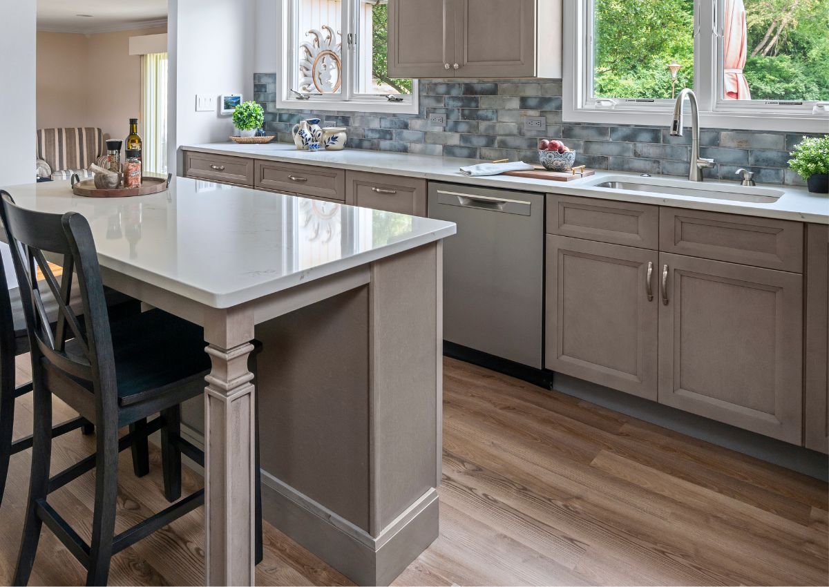 Modern kitchen renovation with light gray cabinets, white countertops, and stainless-steel appliances including a dishwasher. The island has a white countertop with dark wooden stools. The backsplash features blue and gray tiles, and the floor is wood. A window brings in natural light.