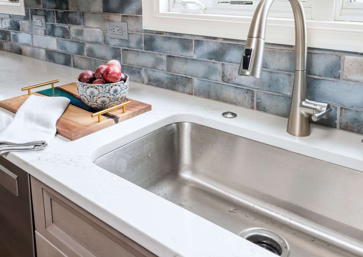 A modern kitchen sink with a tall, curved faucet set in a sleek white countertop. Next to the sink, there's a decorative bowl holding red apples on a wooden cutting board with gold handles. A white towel is placed on the cutting board. The backsplash features blue and gray tiles, perfect for any kitchen renovation.