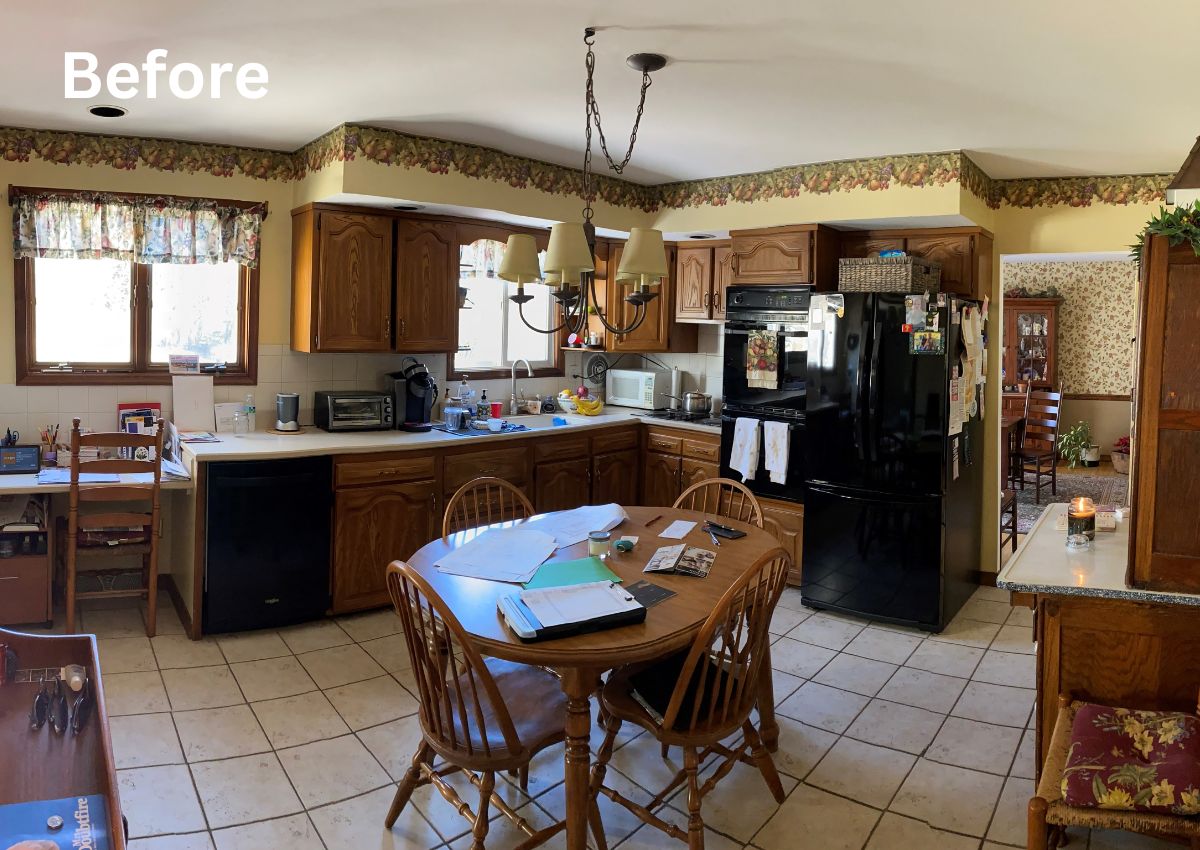 A kitchen with wooden cabinets, a black refrigerator, white dishwasher, microwave, and various items on the countertops. A dining table with chairs is in the center, holding papers and a few other objects. A floral valance is above the windows, and the word "Before" is in the top left corner, highlighting this space as ripe for kitchen renovation.