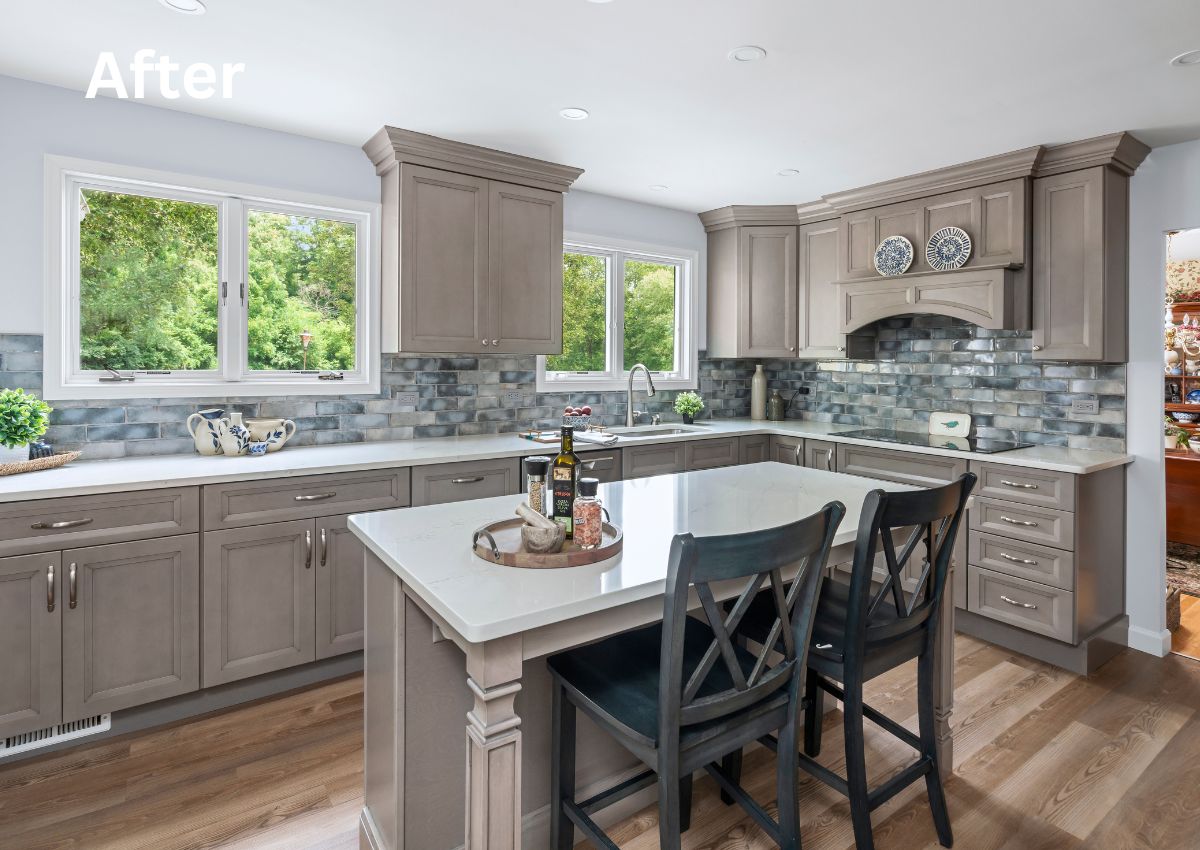 A modern kitchen renovation with light grey cabinets, white countertops, and a tiled backsplash. An island with wine and oil bottles, accompanied by two black chairs, is central. Large windows offer a view of greenery outside. "After" is written in the upper left corner.