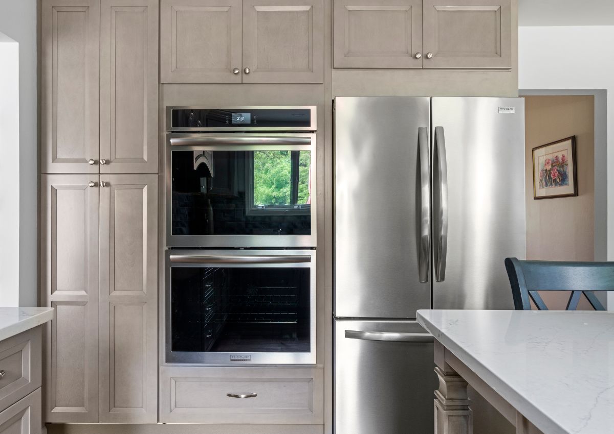 A modern kitchen renovation features light grey cabinetry, a double wall oven, and a stainless steel refrigerator. A bright window reflected in the oven door adds natural light, and a white countertop is partially visible in front. Framed artwork hangs on the wall in the background.