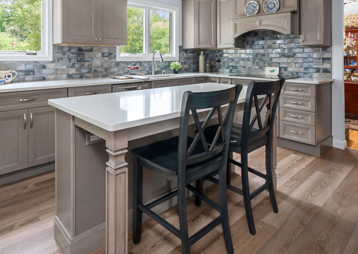 A modern kitchen renovation with light gray cabinets and a matching island. The countertop is white, and the backsplash consists of blue-gray tiles. Two black barstools are placed at the island. The kitchen features hardwood flooring and a few decorative items.