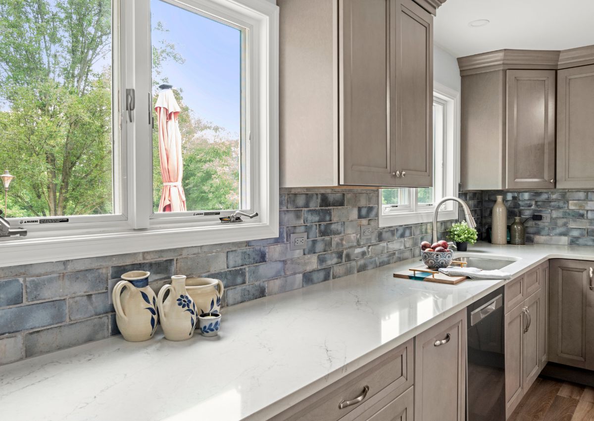 A modern kitchen renovation with light gray cabinets and white marble countertops. The backsplash features rectangular blue-gray tiles. Sunlight streams through the large windows, offering a view of greenery outside. Decorative pitchers are thoughtfully placed on the counter.