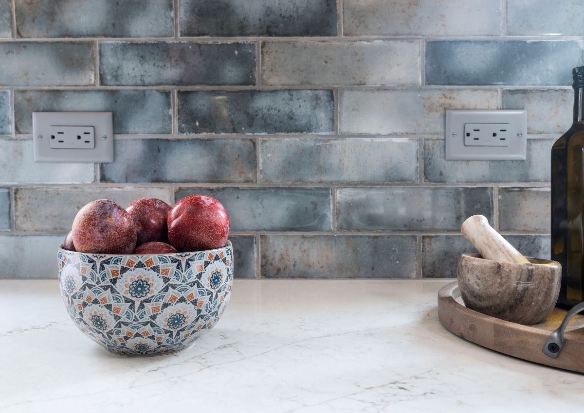 A kitchen counter, fresh from a recent renovation, features a decorative bowl with red pomegranates set against a patterned blue, gray, and brown tile backsplash. Nearby, a mortar and pestle sit on a wooden tray alongside a bottle of olive oil. 