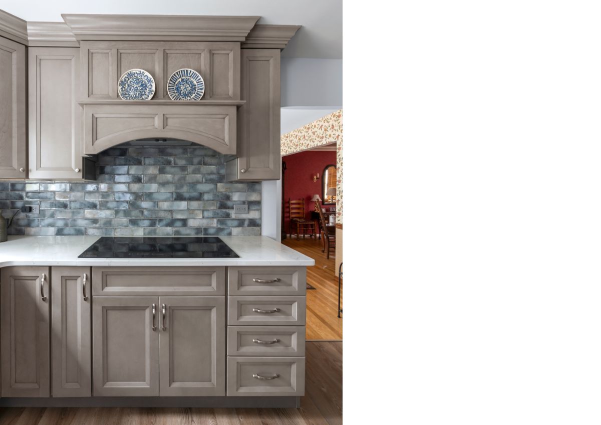 A recent kitchen renovation showcases gray cabinetry, a white countertop, and a backsplash with gray and blue tiles. Decorative plates are displayed in a wooden compartment above the stove. The background reveals a dining area with red wallpaper and wooden flooring.