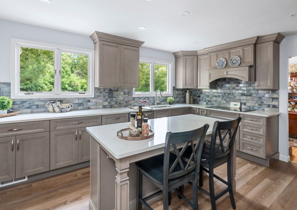 Spacious modern kitchen renovation with gray cabinets, white countertops, and a central island with two dark chairs. A backsplash with shades of gray and blue complements the decor. Large windows provide ample natural light, and various kitchen items adorn the countertops.