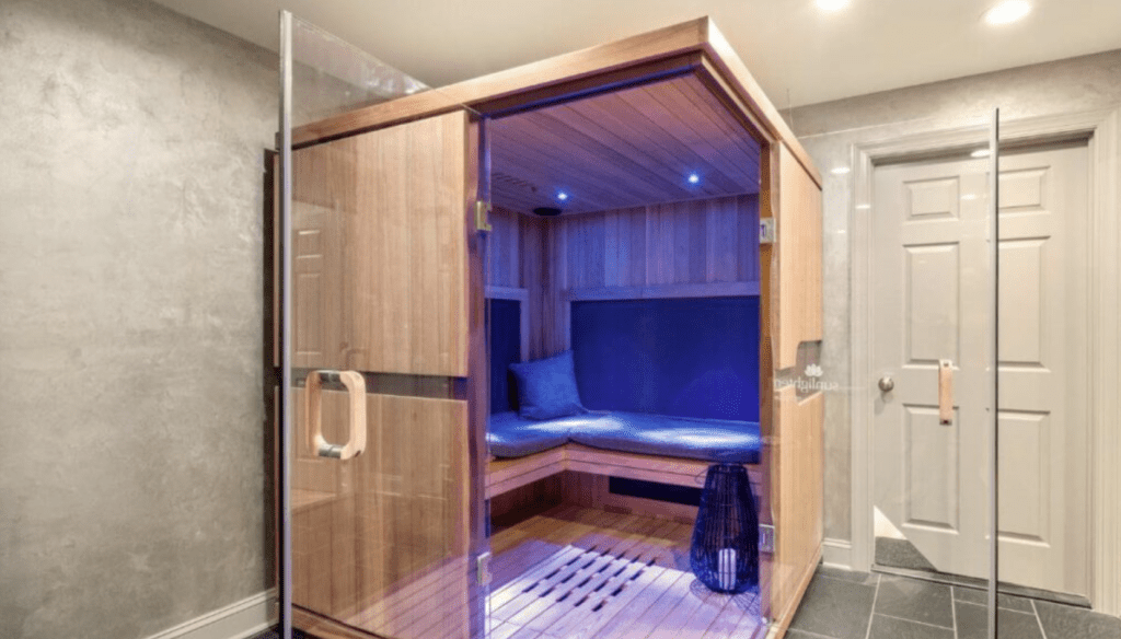 A modern steam sauna room with glass doors, wood paneling, and interior blue LED lighting. The sauna features a built-in bench with cushions and a dark stone floor surrounding it. A closed white door, reminiscent of those seen in high-end kitchen renovations, is visible to the right of the sauna.