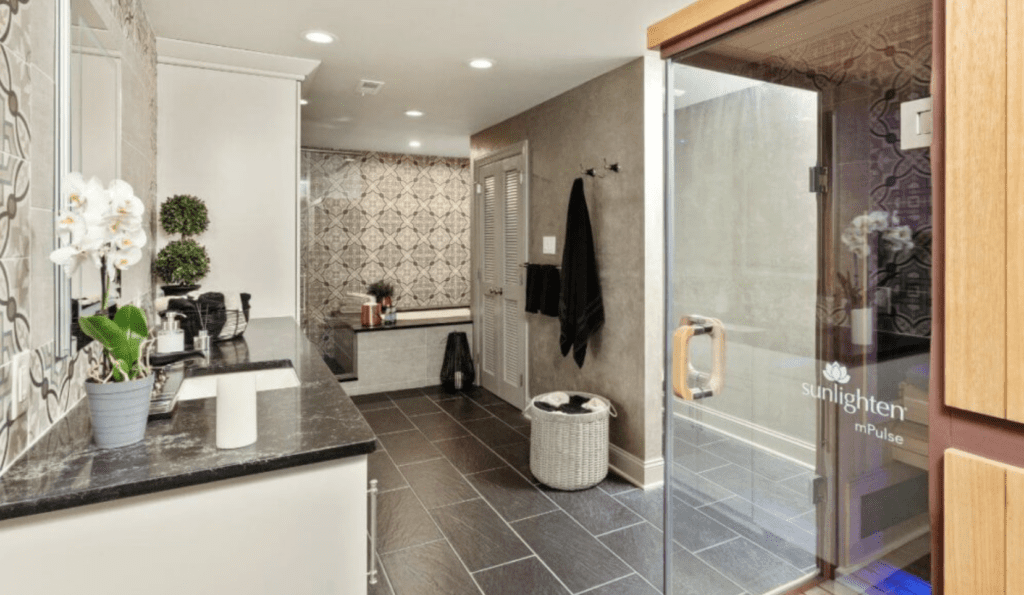 A modern bathroom featuring a black granite countertop with double sinks, a glass-enclosed sauna labeled "Sunlighten mPulse," a large bathtub with surrounding patterned tiles, and a wicker laundry basket reminiscent of those found in stylish kitchen renovations. The room is lit with recessed lighting.